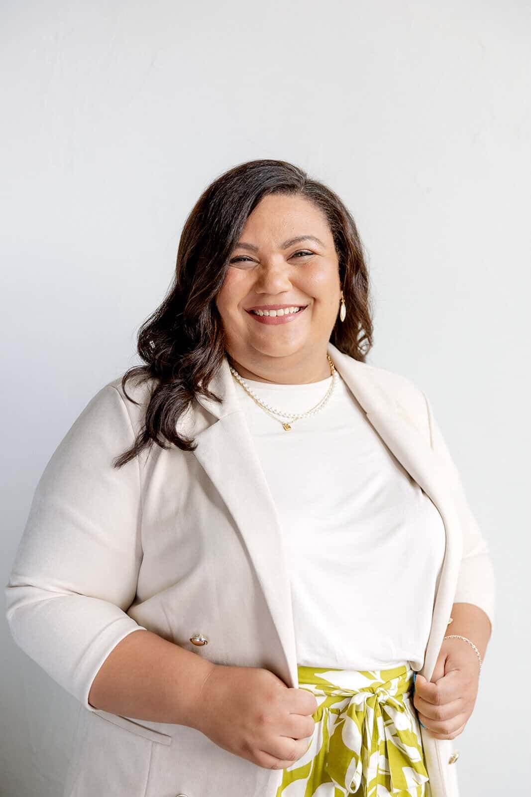 A happy woman in a cream sports coat and white shirt stands smiling against a wall