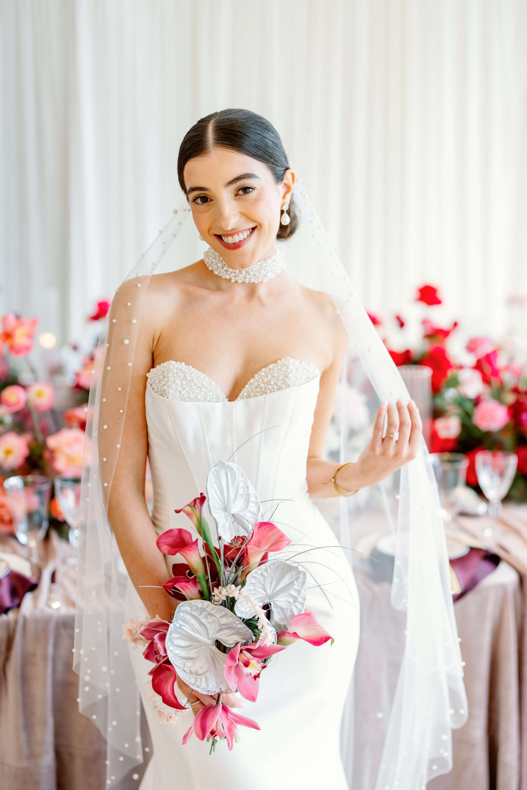 A smiling bride stands in her pearl covered veil holding a pink and white bouquet