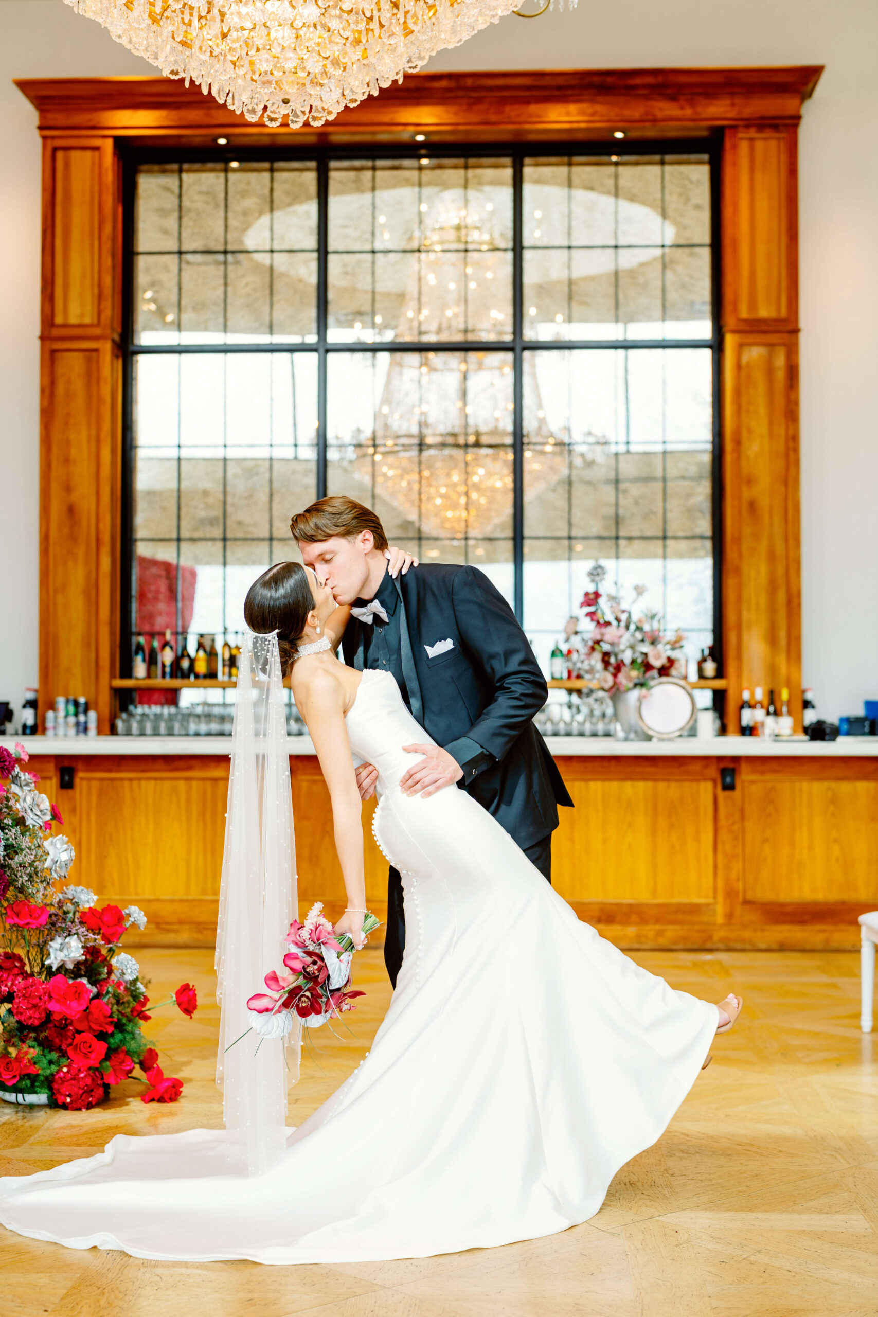 A bride is dipped and kissed by her groom under a crystal chandelier holding a red bouquet next to arrangements planned by Love and Whimsy Events