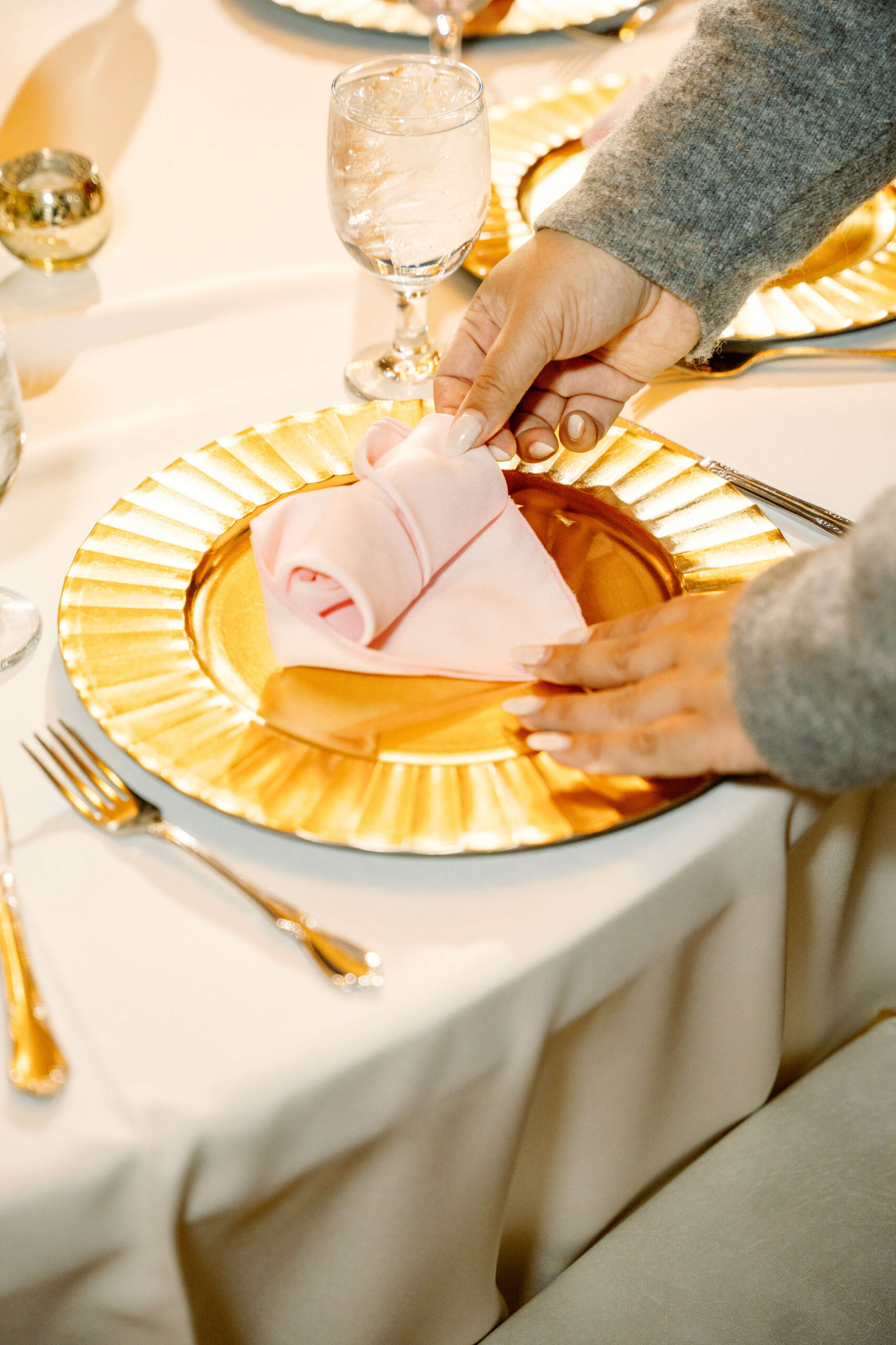 Details of a woman rolling a pink napkin on a gold charger at a wedding reception with Love and Whimsy Events