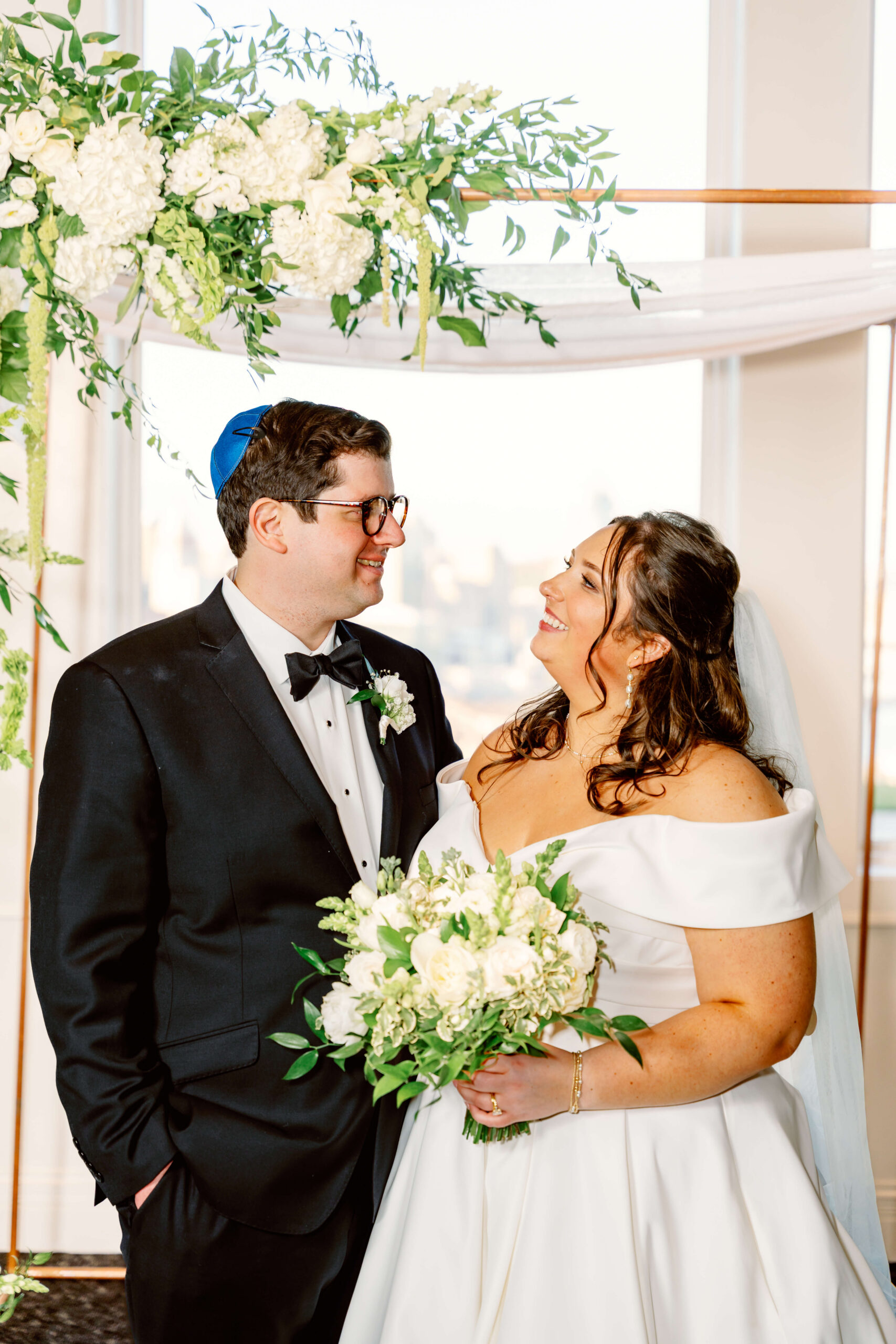 A bride and groom smile at each other under a simple wooden arbor