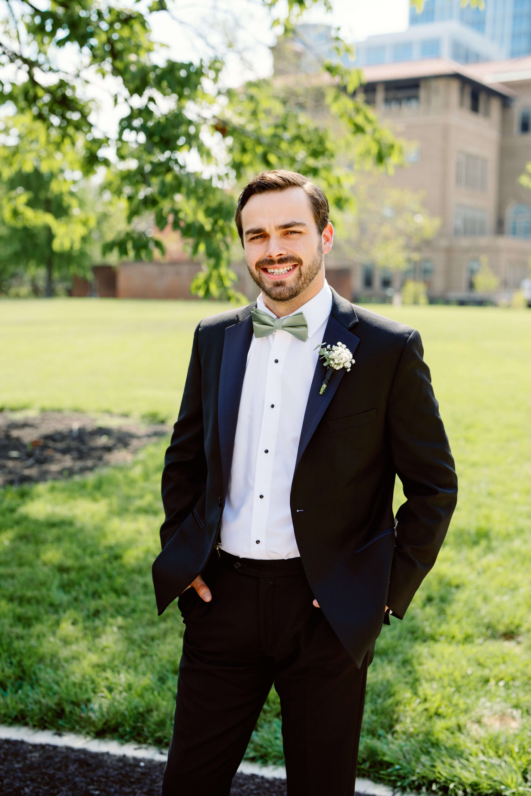 A groom smiles with hands in pockets in a lawn in downtown