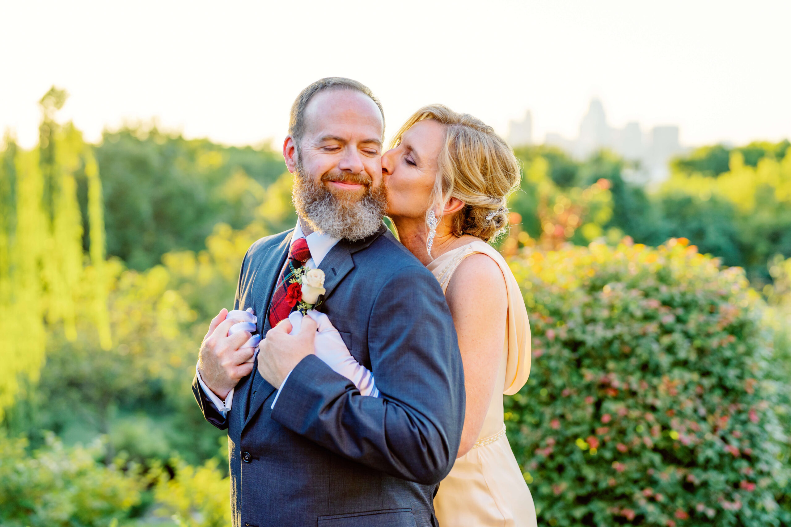 A bride kisses the cheek of her groom while hugging him from behind with gloves on in a garden at sunset