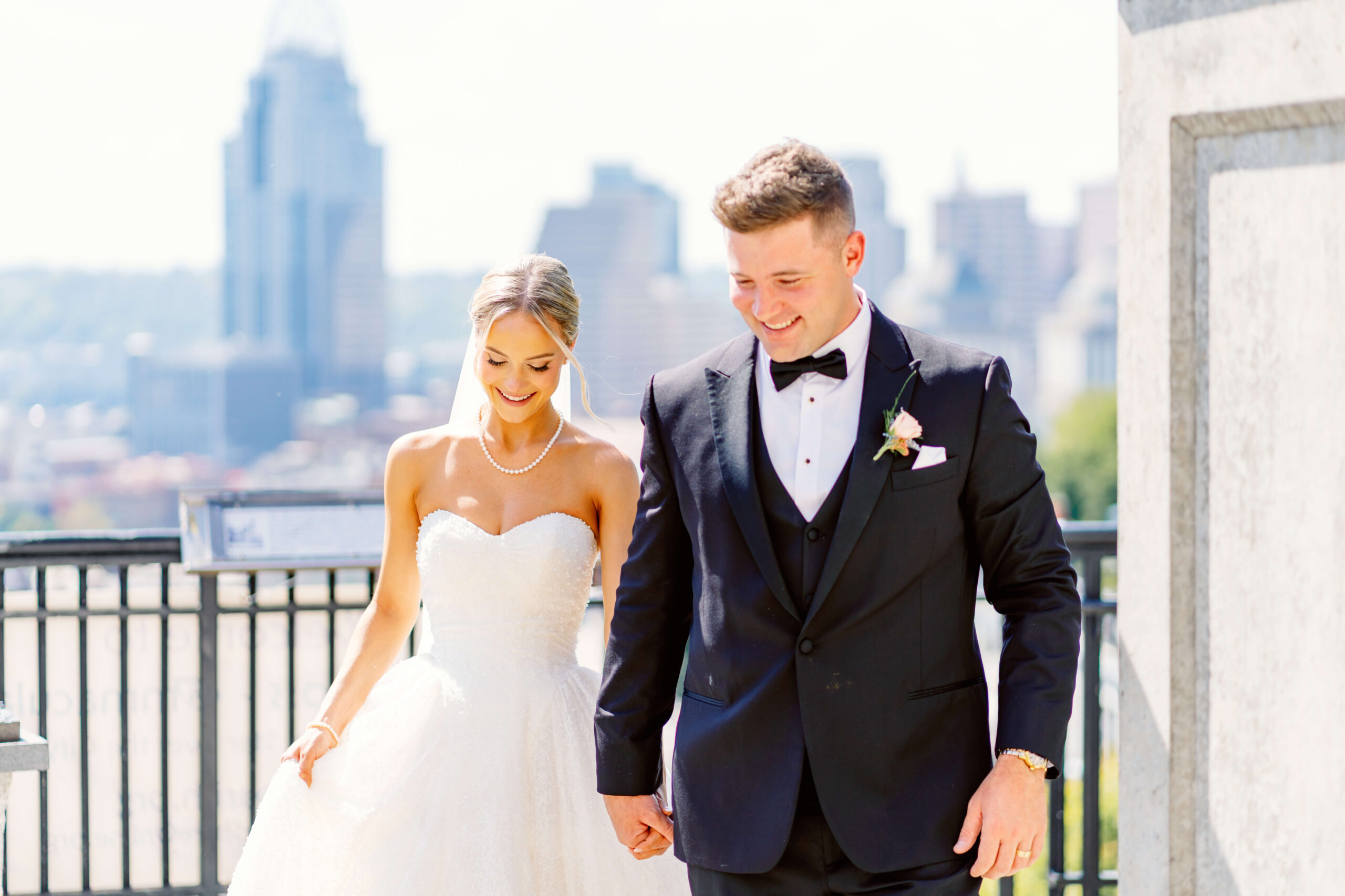 A bride and groom laugh while walking together holding hands on an overlook with the skyline across the river