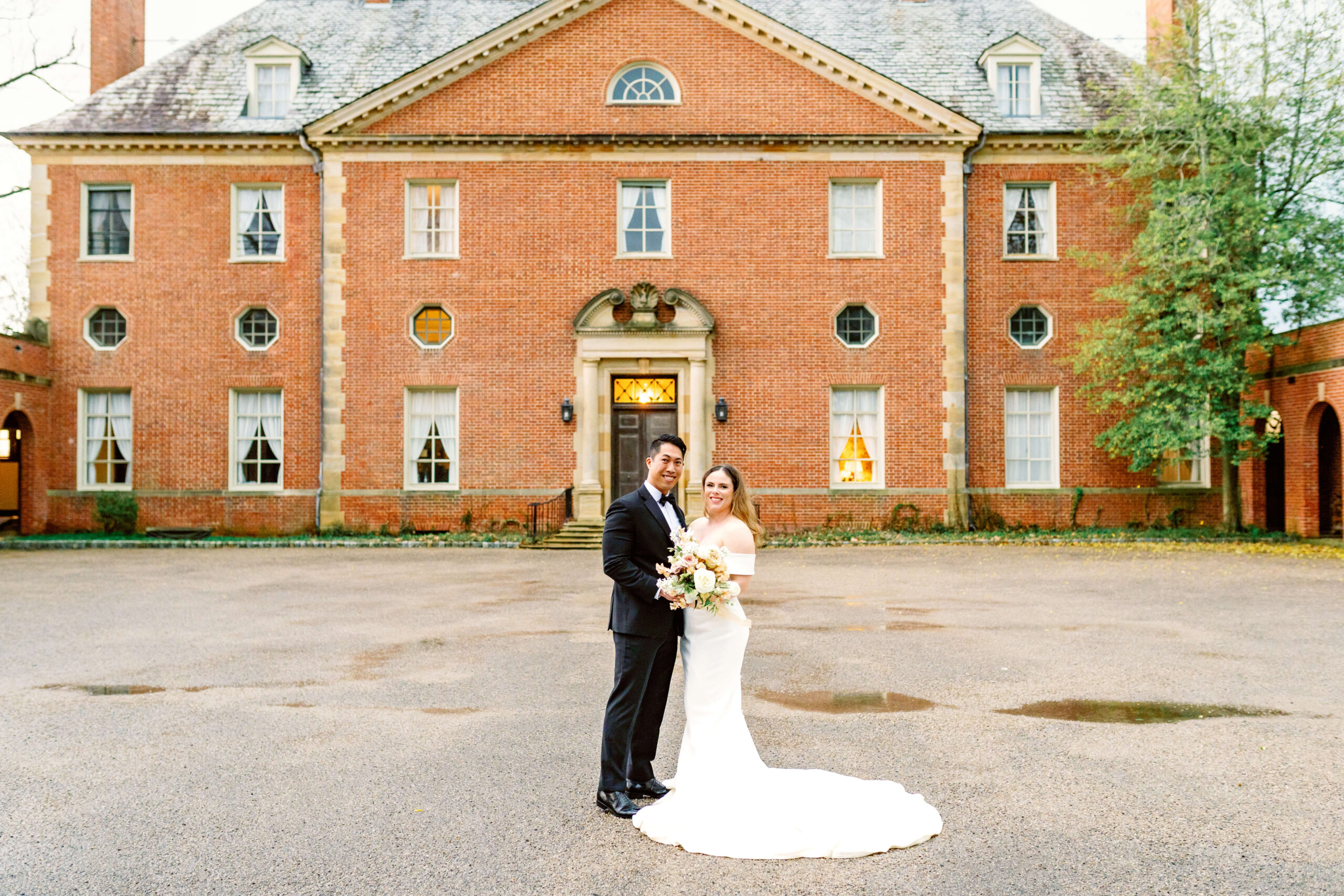 A bride and groom stand in front of one of the estate wedding venues in cincinnati smiling and holding a large bouquet