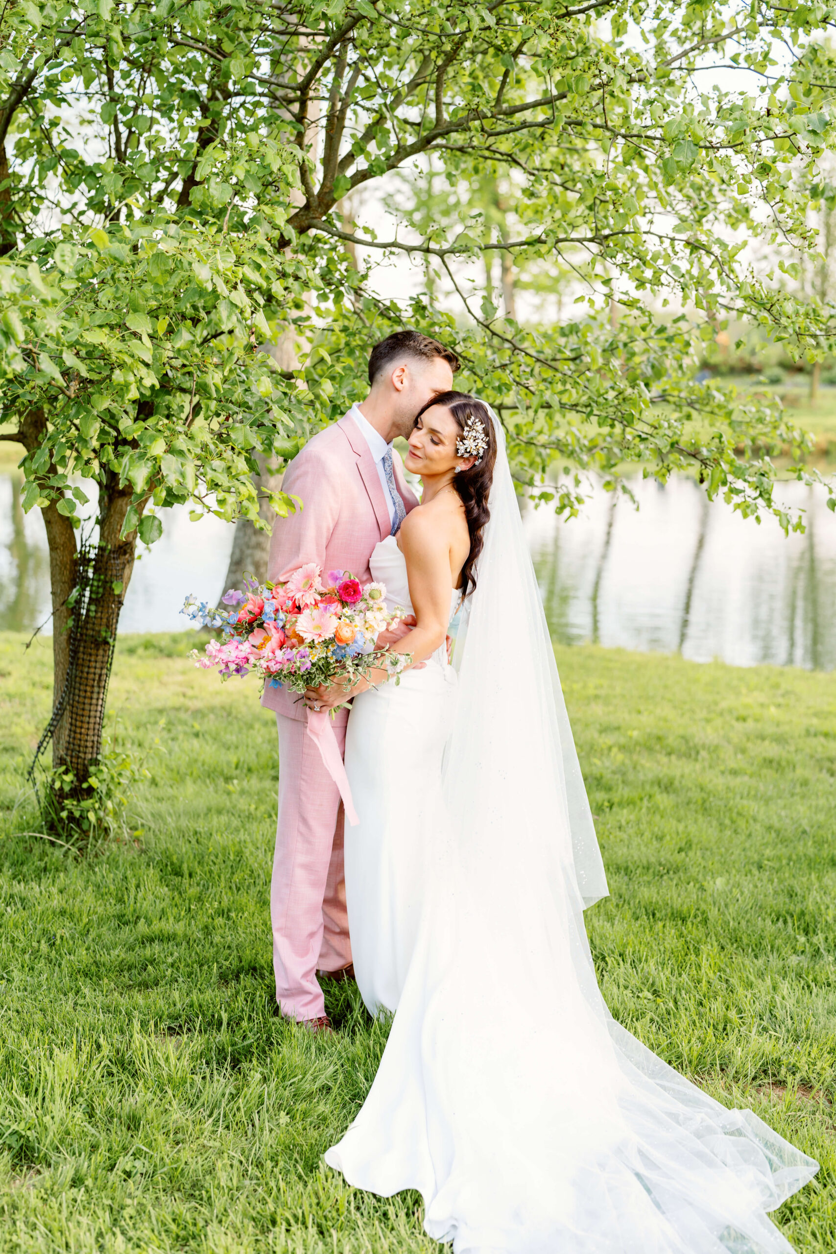 Newlyweds snuggle under a tree by a pond with smiles and holding a bride bouquet