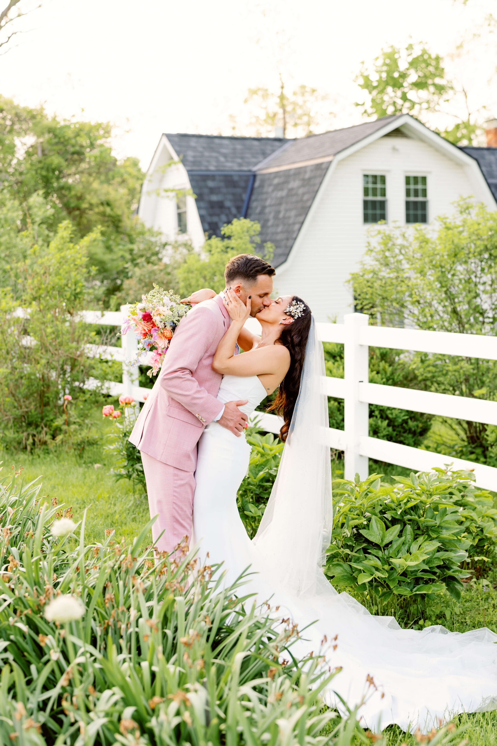 A groom in a pink suit kisses his bride in a garden along a white fence at one of the unique wedding venues in cincinnati