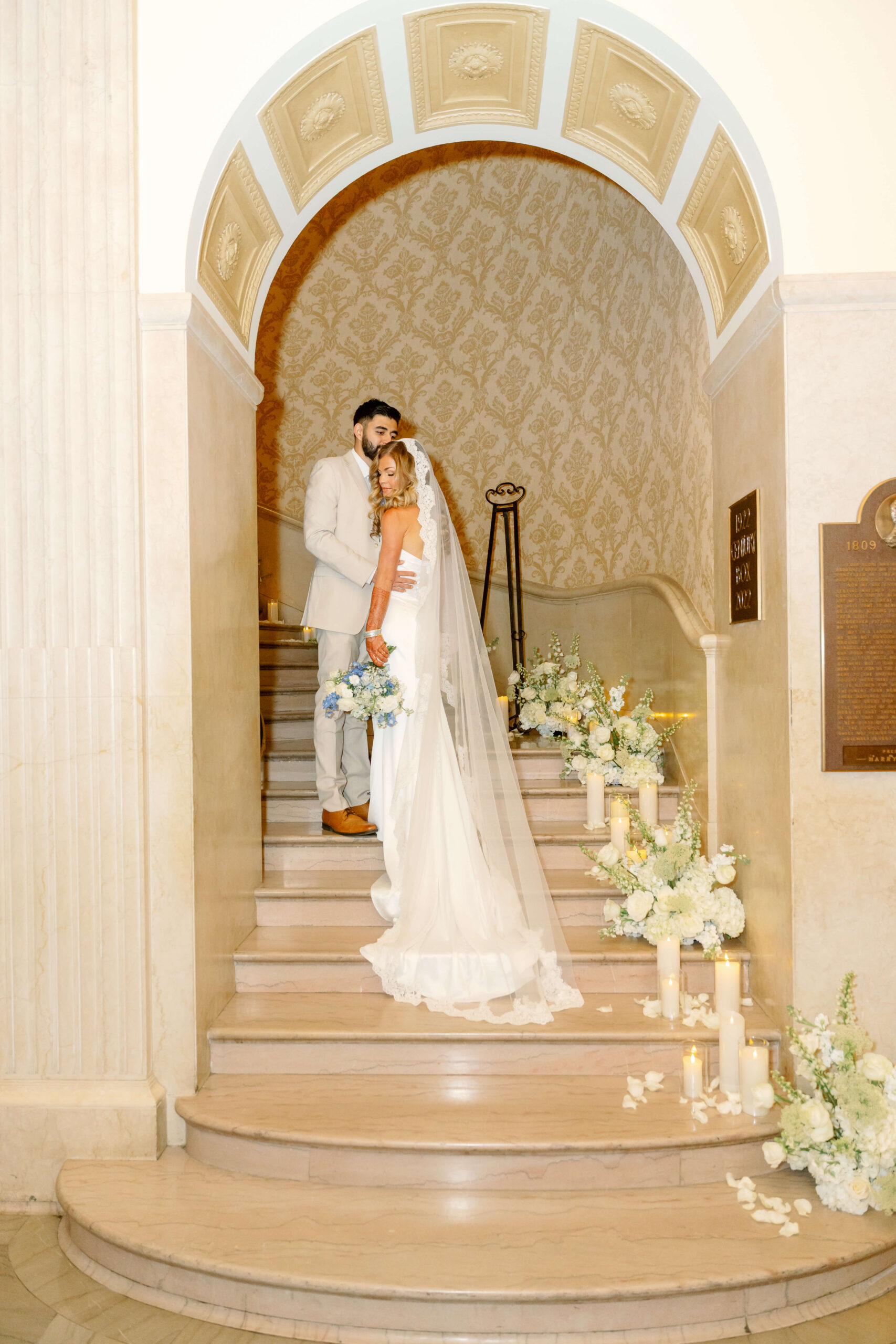 A bride snuggles her groom in a tan suit on a grand marble staircase lined with candles and flowers