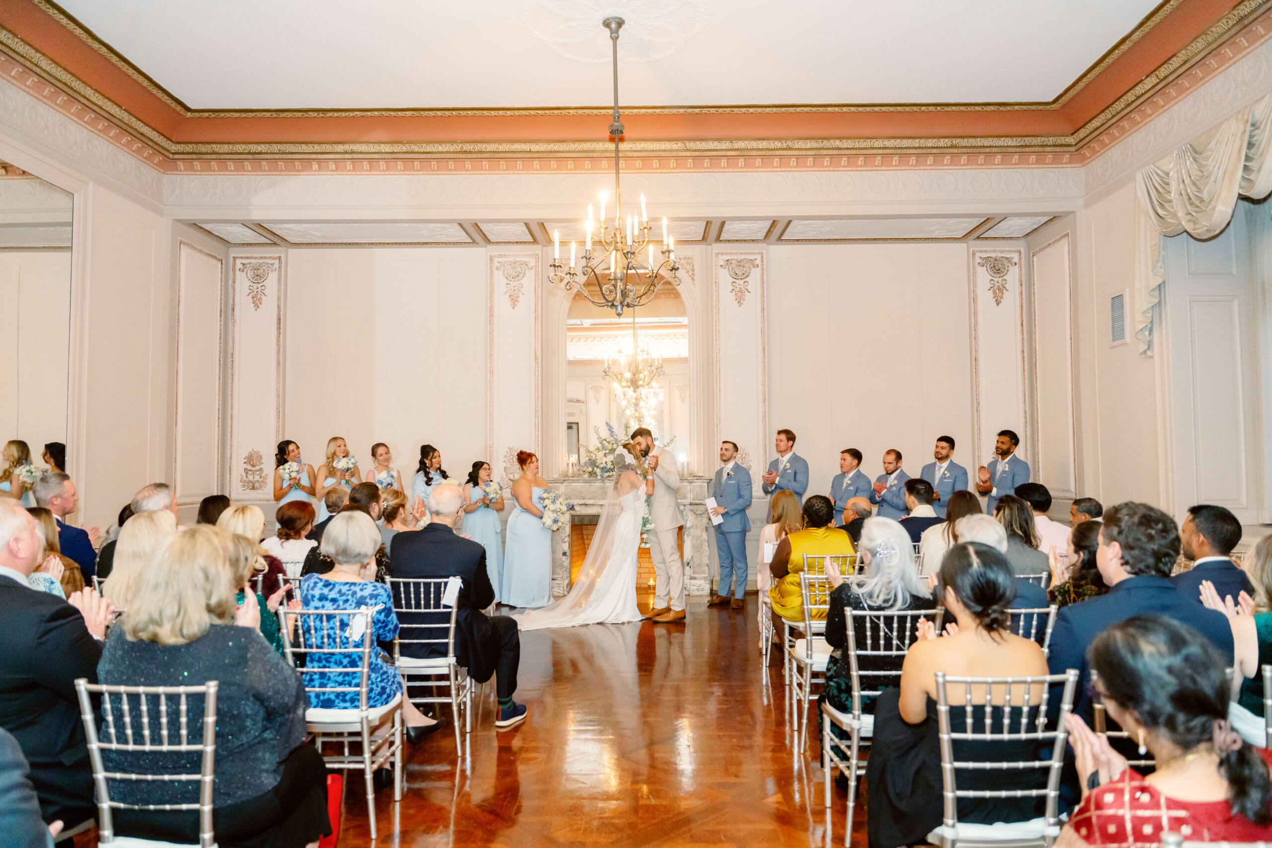 Newlyweds hold hands and kiss to end their indoor ceremony under a chandelier at one of the unique wedding venues in cincinnati as guests clap