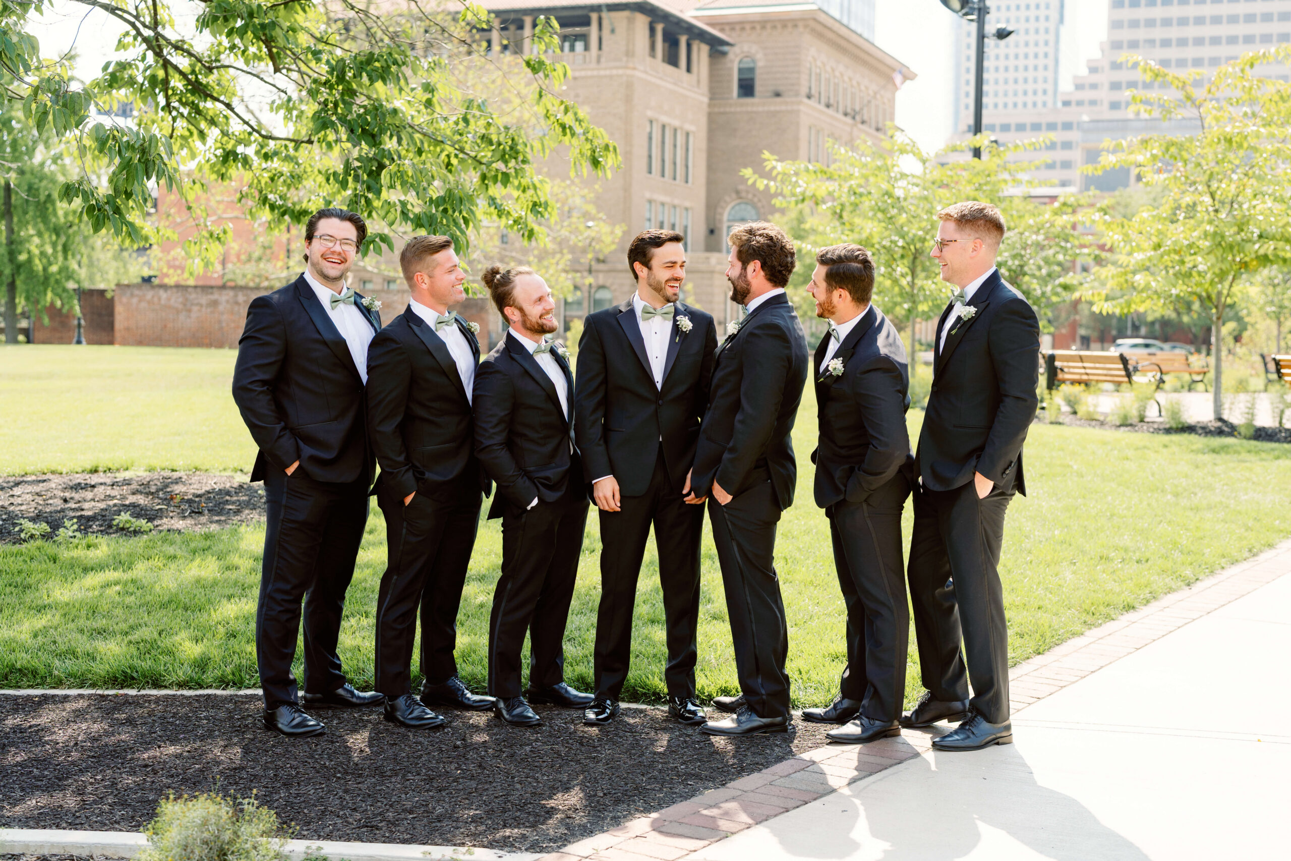 A groom in a black suit laughs with his groomsmen in a park lawn with hands in pockets