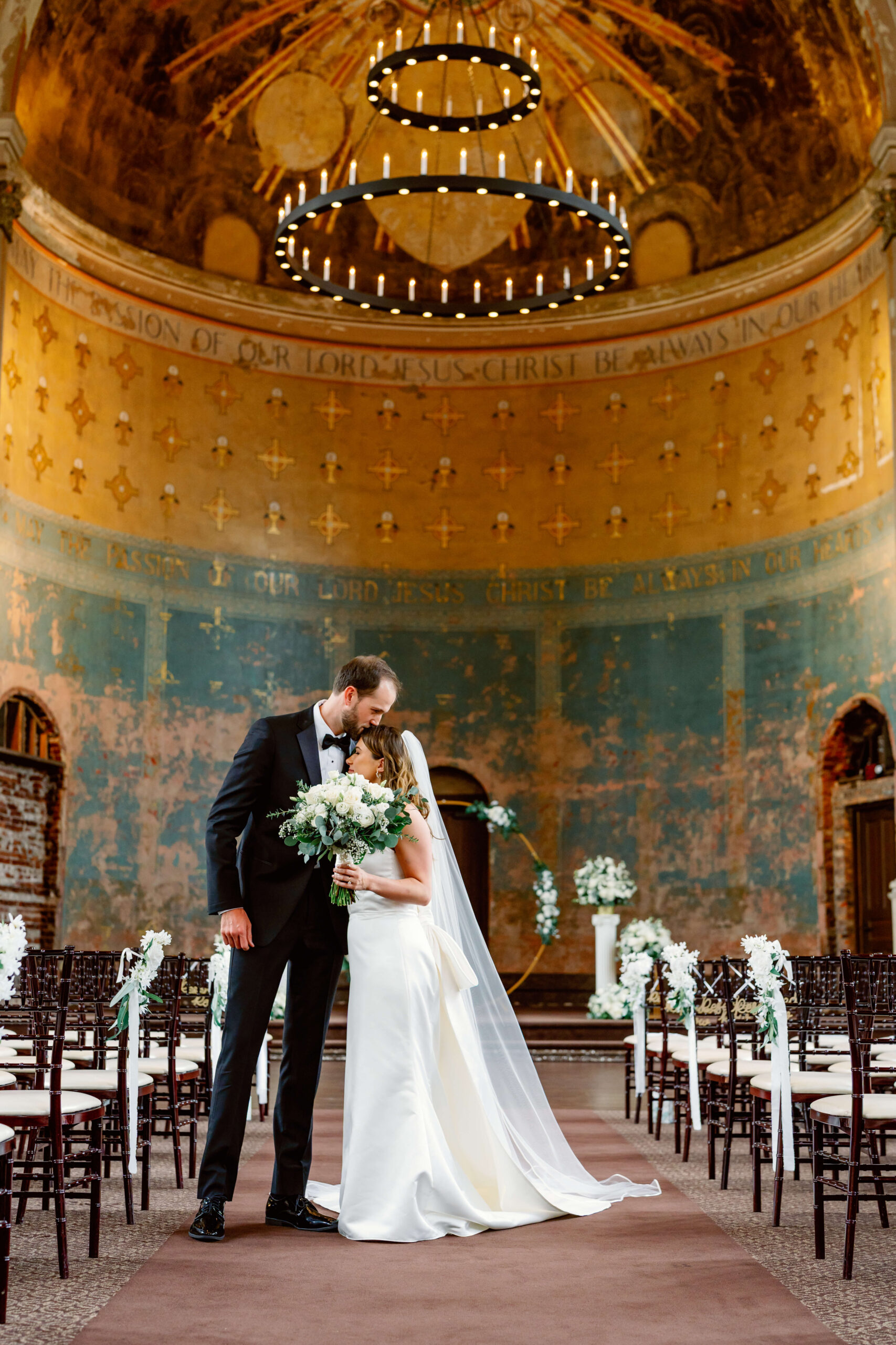 A groom leans over to kiss his bride on the head in an old chapel at one of the unique wedding venues in cincinnati