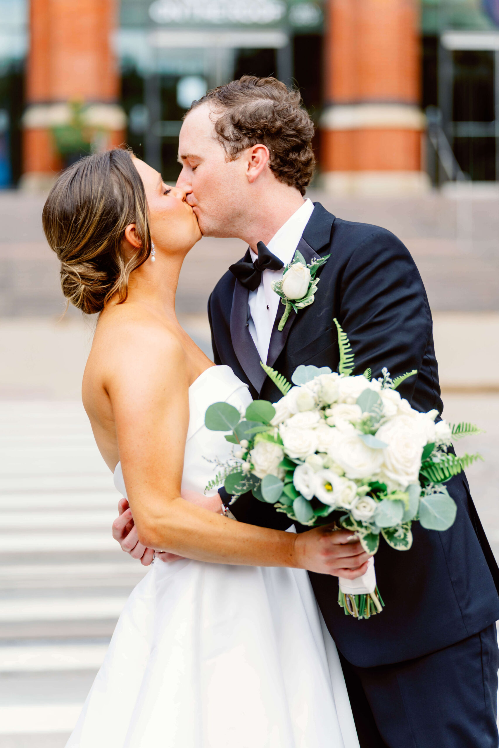 A bride and groom kiss on a crosswalk while holding a large white bouquet